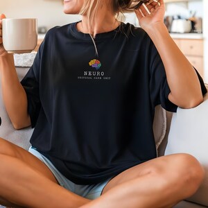 May include: Black t-shirt with a colorful brain graphic and the text "NEURO CRITICAL CARE UNIT". The shirt is a crew neck style and appears to be made of a soft material. A black circle with the word "Black" is in the lower left corner.