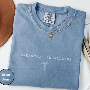 May include: A blue jean colored short-sleeve t-shirt with a crew neckline. The chest features the white text EMERGENCY DEPARTMENT above a white caduceus symbol. A gold-toned necklace with a small circular pendant rests on the fabric.