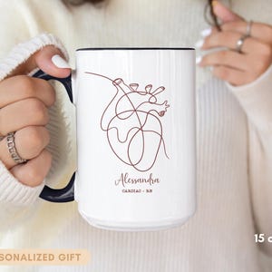 May include: White ceramic mug with a black handle and rim, featuring a minimalist line drawing of a heart. The mug is personalized with a name and the text "Cardiac - RN". The mug is 15 oz.