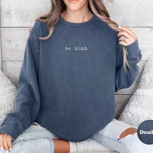 May include: A dark blue sweatshirt with the text "be kind." embroidered in white on the front. The sweatshirt is worn by a person with long brown hair and is sitting on a white fluffy surface.
