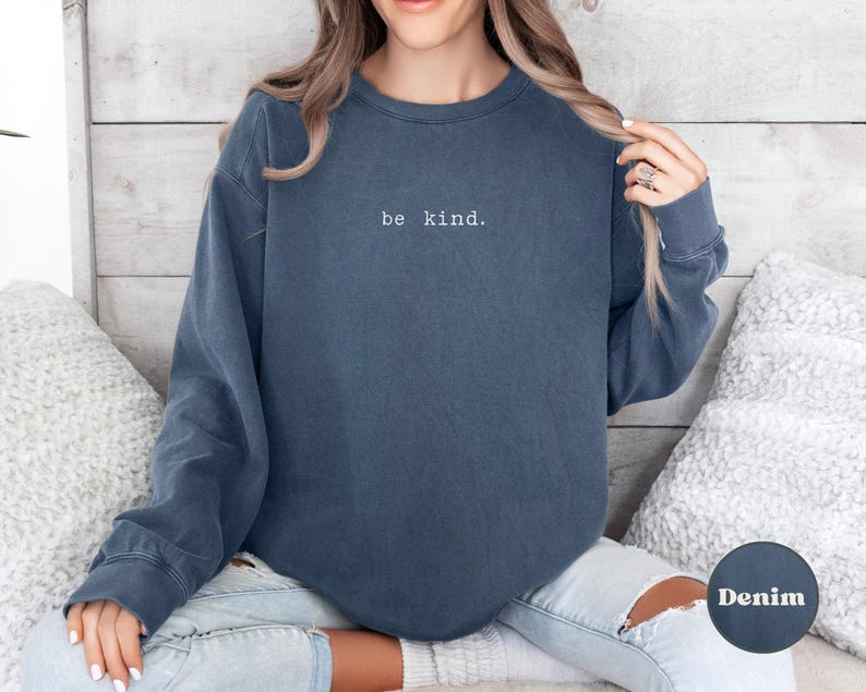 May include: A dark blue sweatshirt with the text "be kind." embroidered in white on the front. The sweatshirt is worn by a person with long brown hair and is sitting on a white fluffy surface.