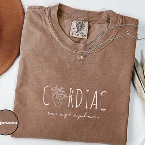 May include: Espresso-colored t-shirt with the words "CARDIAC sonographer" in white script. The design includes a stylized heart with floral elements. A silver necklace with a pendant rests on the shirt.