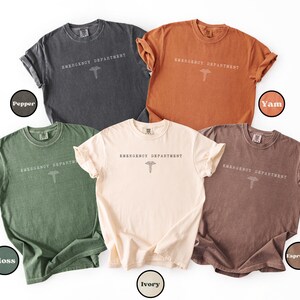 May include: Five short-sleeve crew neck t-shirts in pepper, yam, moss, ivory, and espresso colors. Each shirt features the text EMERGENCY DEPARTMENT printed in a simple font above a caduceus symbol centered on the chest area of the garment.