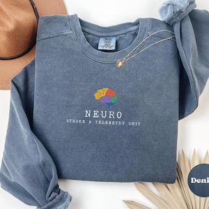 May include: Denim blue sweatshirt with the word "NEURO" and a colorful brain graphic. Below the word "NEURO" is the text "STROKE & TELEMETRY UNIT". A gold necklace is draped over the sweatshirt.