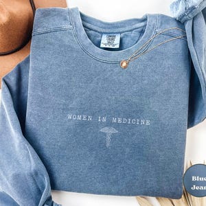 Puede incluir: Una sudadera de mezclilla azul con el texto "Women in Medicine" y un s&iacute;mbolo de caduceo.