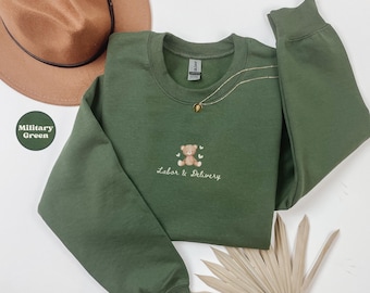 Op de afbeelding: Militairgroen sweatshirt met een ronde hals, geborduurd met een teddybeer en de woorden "Labor & Delivery". Een gouden ketting is zichtbaar. Een bruine hoed staat op de achtergrond. Het sweatshirt is groen.