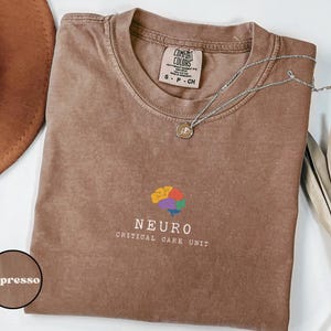 May include: Espresso-colored t-shirt with a crew neck and short sleeves. The shirt features a colorful brain graphic and the words "NEURO CRITICAL CARE UNIT" in white. A silver necklace with a pendant rests on the shirt.
