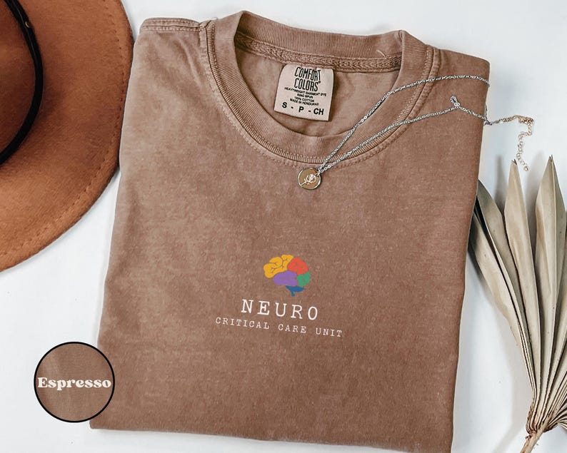 May include: Espresso-colored t-shirt with a crew neck and short sleeves. The shirt features a colorful brain graphic and the words "NEURO CRITICAL CARE UNIT" in white. A silver necklace with a pendant rests on the shirt.