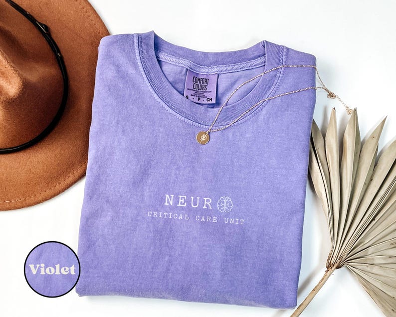 May include: A violet-colored t-shirt with the text "NEUR CRITICAL CARE UNIT" in white. A gold necklace with a pendant rests on the shirt. A brown hat and dried palm leaves are also in the image.