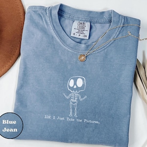 May include: A blue jean Comfort Colors t-shirt with a white skeleton graphic and the text "IDK I Just Take the Pictures...". A gold necklace with a pendant is draped on the shirt.
