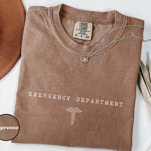May include: An espresso-colored short-sleeve t-shirt with a crew neckline. The chest features the white text EMERGENCY DEPARTMENT above a white caduceus symbol. The inner neck label reads Comfort Colors, 100% cotton, size S-P-CH.