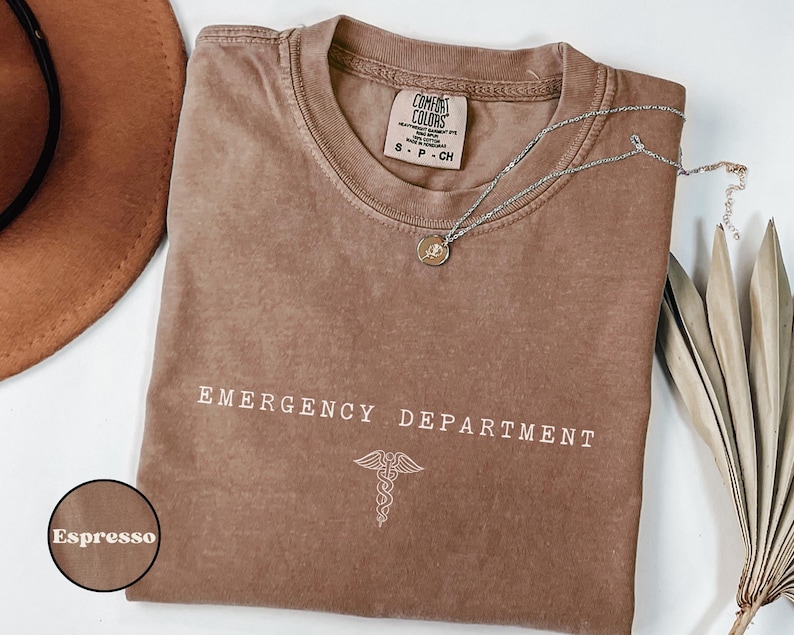 May include: An espresso-colored short-sleeve t-shirt with a crew neckline. The chest features the white text EMERGENCY DEPARTMENT above a white caduceus symbol. The inner neck label reads Comfort Colors, 100% cotton, size S-P-CH.