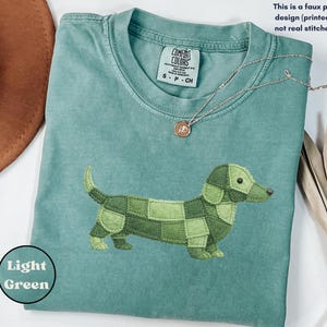 May include: A light green Comfort Colors t-shirt featuring a patchwork dachshund design in shades of green. The shirt has a crew neck and short sleeves. The text "Light Green" is in a circle on the bottom left.