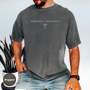 May include: A charcoal grey short-sleeve t-shirt featuring the text EMERGENCY DEPARTMENT printed in white on the chest, positioned above a white line-art caduceus symbol. The shirt has a crew neckline and a relaxed, loose-fitting silhouette.