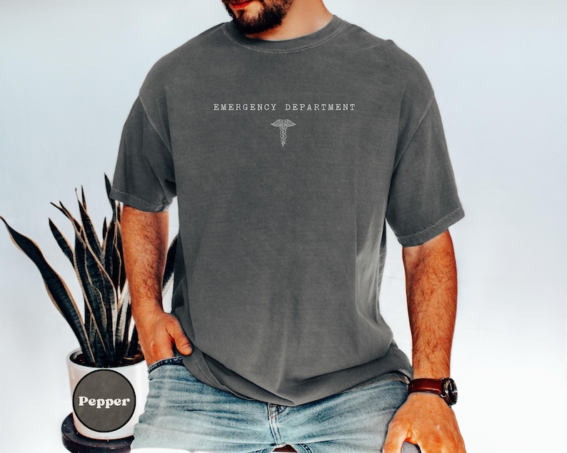 May include: A charcoal grey short-sleeve t-shirt featuring the text EMERGENCY DEPARTMENT printed in white on the chest, positioned above a white line-art caduceus symbol. The shirt has a crew neckline and a relaxed, loose-fitting silhouette.