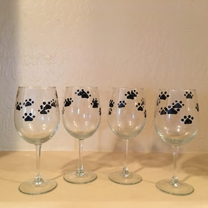 May include: Four clear wine glasses with black paw print designs. The glasses are arranged in a row, each with a long stem and a rounded bowl. The paw prints are evenly spaced across the surface of each glass.
