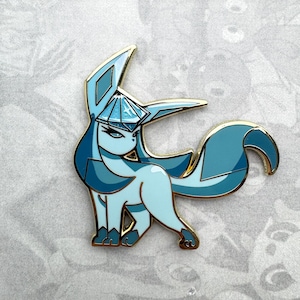 May include: A blue and gold enamel pin featuring a stylized cartoon of a mythical creature with a crown. The creature has a long, pointed tail and a large, pointed ear.