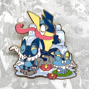 May include: A blue, yellow, and pink enamel pin featuring three Pokemon characters: a blue cat-like Pokemon with a white ruff, a yellow bird-like Pokemon with a pink scarf, and a blue frog-like Pokemon. The Pokemon are sitting on a blue and white background with a waterfall and pink flowers.