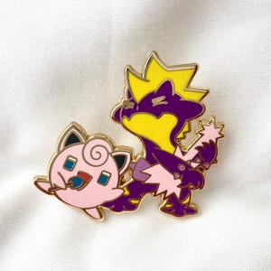 May include: A gold-coloured enamel pin featuring a purple and yellow Pokemon character holding a pink Pokemon character. The pin is a cartoon style illustration.