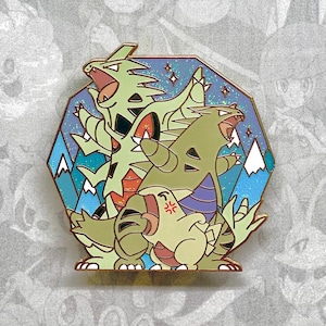 Puede incluir: Un pin de esmalte verde, dorado y azul con tres Pokémon: un Tyrantrum, un Tyrunt y un Amaura. El pin tiene forma de hexágono y un fondo brillante.
