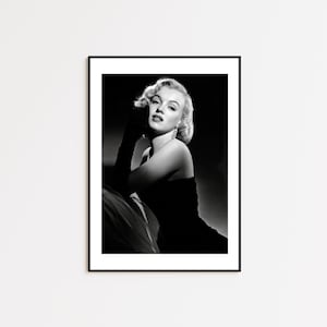 May include: Black and white framed print of a woman with blonde hair, wearing a strapless dress and long gloves. She is leaning on her arm, looking towards the viewer. The print is in a black frame.