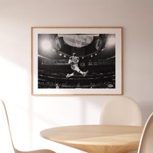 Puede incluir: Fotograf&iacute;a en blanco y negro de un jugador de baloncesto en el aire, a punto de hacer un mate. El jugador lleva una camiseta con el n&uacute;mero 23. La foto fue tomada en un estadio con una multitud de personas en el fondo.
