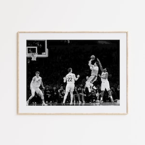 May include: Black and white framed photograph of a basketball game in action. A player in mid-air attempts a shot, while two opposing players jump to block. The crowd is visible in the background.