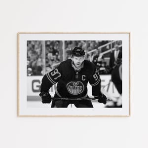 May include: Black and white photo of a hockey player wearing a black and white jersey with the number 97 and the word "Oilers" on the front. The player is holding a hockey stick and is looking at the camera.