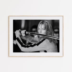 Op de afbeelding: Zwart-wit ingelijste print met een vrouw die een katana-zwaard vasthoudt. Ze draagt een geel en zwart trainingspak. De afbeelding is een portret, waarbij de vrouw opzij kijkt en het zwaard vasthoudt.