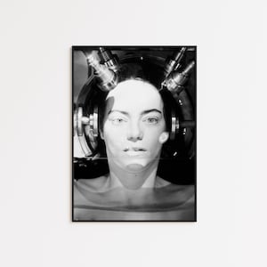 May include: Black and white photo of a person's face with a device attached to their head. The device has multiple metal arms and appears to be a medical or scientific instrument.