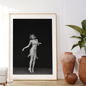 May include: Black and white photograph of a woman wearing a fringed dress, posing in a studio setting.