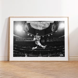 Puede incluir: Fotograf&iacute;a en blanco y negro de un jugador de baloncesto en el aire, a punto de hacer un mate. El jugador lleva una camiseta con el n&uacute;mero "23". La foto fue tomada en un estadio con una gran multitud de fondo.