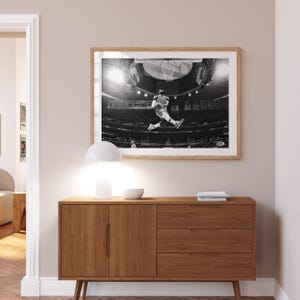Puede incluir: Fotograf&iacute;a en blanco y negro de un jugador de baloncesto en el aire, a punto de hacer un mate. El jugador lleva una camiseta y pantalones cortos. La foto est&aacute; enmarcada y colgada en una pared sobre una c&oacute;moda de madera.