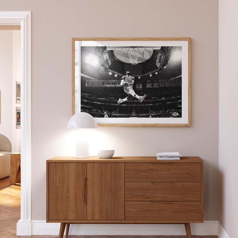 Puede incluir: Fotograf&iacute;a en blanco y negro de un jugador de baloncesto en el aire, a punto de hacer un mate. El jugador lleva una camiseta y pantalones cortos. La foto est&aacute; enmarcada y colgada en una pared sobre una c&oacute;moda de madera.