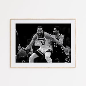 May include: Black and white framed photograph of a basketball game. Two players are in action, one in a white jersey with "WOLVES 21" and the other in a black jersey with "26". A basketball is visible.