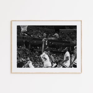 以下が含まれることがあります： バスケットボール選手が空中でダンクシュートを決めようとしている白黒写真。選手は背番号12のユニフォームを着ています。写真はスタンドから撮影されており、背景には観客が見えます。