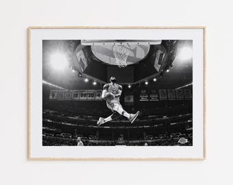 May include: Black and white photograph of a basketball player in mid-air, about to dunk the ball. The player is wearing a jersey with the number 23 on it. The photo was taken in a stadium with a crowd in the background.