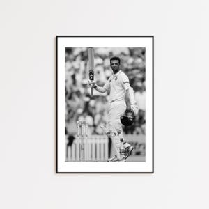 May include: A black and white photo of a cricket player holding a bat and wearing a white uniform. The player is standing on a cricket field with a crowd of people in the background.