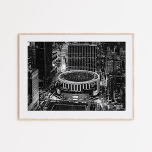 May include: Black and white photograph of Madison Square Garden in New York City, framed in a light wood. The arena is circular, surrounded by tall buildings and city lights. The image is a high-angle shot.