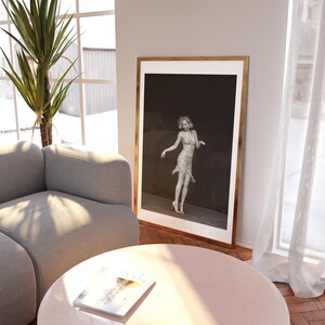 May include: A black and white photograph of a woman in a vintage-style dress, posing in a studio setting. The photo is framed in a gold frame and hanging on a wall.