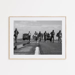 May include: Black and white framed photograph of a group of people walking down a road. Military vehicles are visible in the background. The image is a medium shot, capturing the subjects in motion.