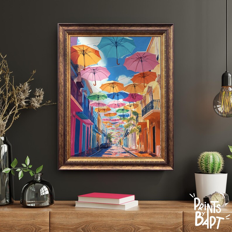 May include: A colourful painting of a street lined with buildings, with many colourful umbrellas hanging overhead. The umbrellas are in shades of pink, yellow, blue, green, and orange. The buildings are painted in a variety of colours, including yellow, orange, red, and blue.