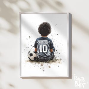 Peut inclure: Une illustration aquarelle d'un jeune portant un maillot de football avec le nom "KYLIAN" et le numéro "10" au dos. La personne est assise avec un ballon de football devant elle, sur un fond blanc avec des éclaboussures grises et brunes.