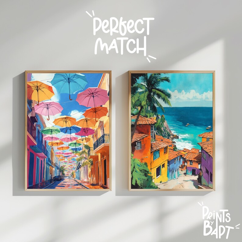 May include: Two framed paintings of colourful cityscapes. The left painting shows a street lined with colourful buildings and a row of colourful umbrellas overhead. The right painting shows a colourful cityscape with a view of the ocean.