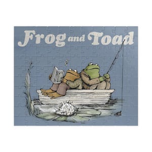 May include: A jigsaw puzzle featuring the text "Frog and Toad" in white letters. The puzzle depicts two anthropomorphic frogs in a boat, one reading a book and the other fishing. The background is a light blue.