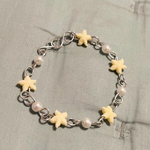 May include: A silver chain bracelet featuring alternating pale yellow starfish charms and white pearl beads. The bracelet is secured with a silver clasp and rests on a light gray surface.