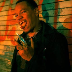 May include: A person with fangs and a spiked ring, wearing a black shirt and a spiked necklace. The image has a dark, moody aesthetic with the text "DEADFANGS" in red at the bottom.