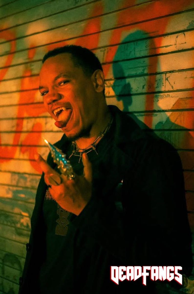 May include: A person with fangs and a spiked ring, wearing a black shirt and a spiked necklace. The image has a dark, moody aesthetic with the text "DEADFANGS" in red at the bottom.