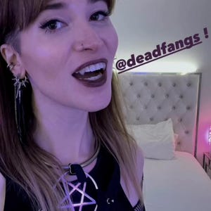 May include: A person with dark lipstick and fangs is wearing a black top with a pentagram pendant. The image includes the text "@deadfangs" and "DEADFANGS" in a stylized font. The person is smiling.