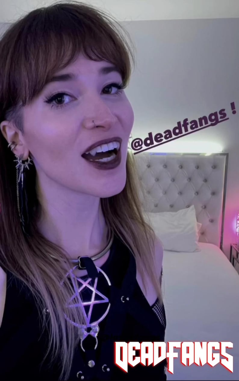 May include: A person with dark lipstick and fangs is wearing a black top with a pentagram pendant. The image includes the text "@deadfangs" and "DEADFANGS" in a stylized font. The person is smiling.